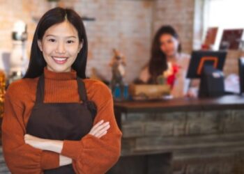 Business owner smiling, as first employee hired completes tasks behind cash register