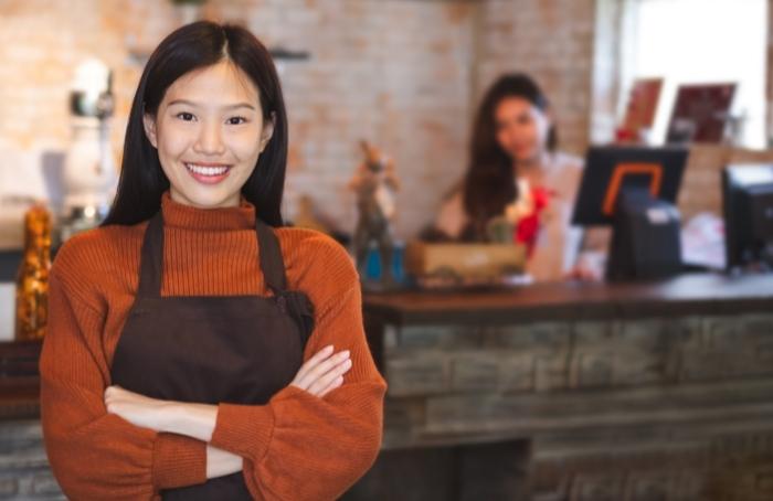 Business owner smiling, as first employee hired completes tasks behind cash register
